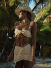 Woman in a beige bikini and hat standing in a tropical setting with palm trees.