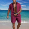 Man in a maroon shirt and shorts standing on a beach with blue water and sky.