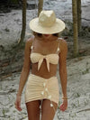 Woman in a beige bikini and hat walking on a sandy path with trees in the background