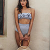 Woman wearing a blue and white striped bikini holding a woven bag against a beige wall.