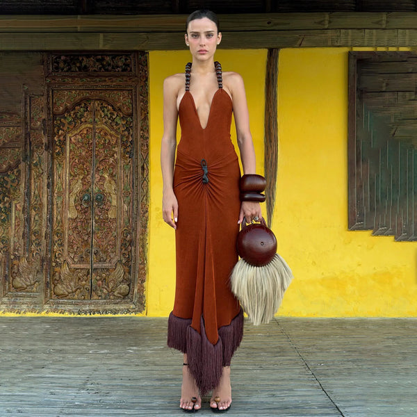 Model wearing a brown dress with fringe details, holding a matching handbag, against a yellow wall.
