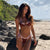 Woman in a red bikini standing on a rocky beach with greenery and ocean in the background